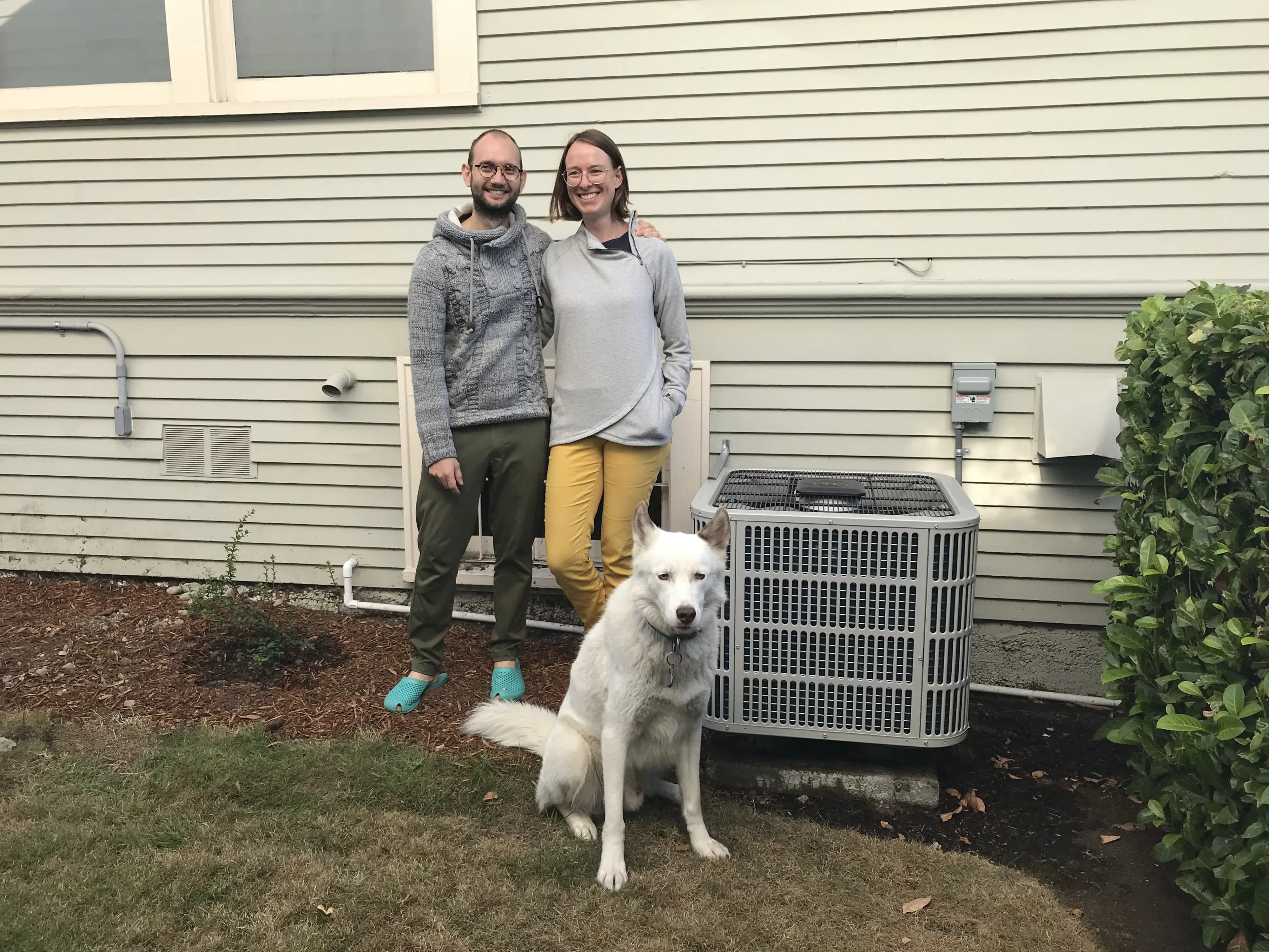 Smiling couple and big fluffy white dog standing next to a heat pump